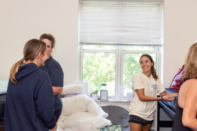 Four people chat in a bright, cozy room with a large window. A young woman in a white T-shirt smiles while arranging items on a desk—showing the welcoming atmosphere of West Chester University housing, complete with pillows, blankets, and soft natural light.