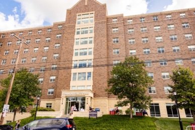 A large brick and stone dormitory building with multiple windows and trees in front serves as West Chester University housing. People gather near the entrance, cars line the street, and a speed limit sign stands in the foreground.