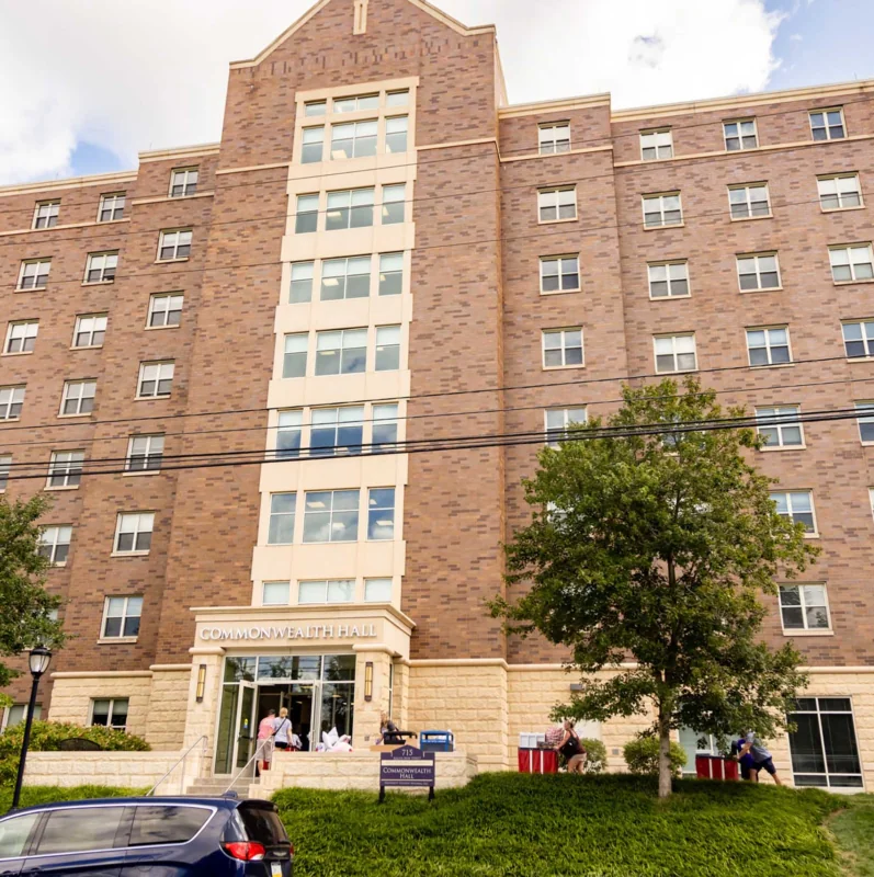 A large brick and stone dormitory building with multiple windows and trees in front serves as West Chester University housing. People gather near the entrance, cars line the street, and a speed limit sign stands in the foreground.