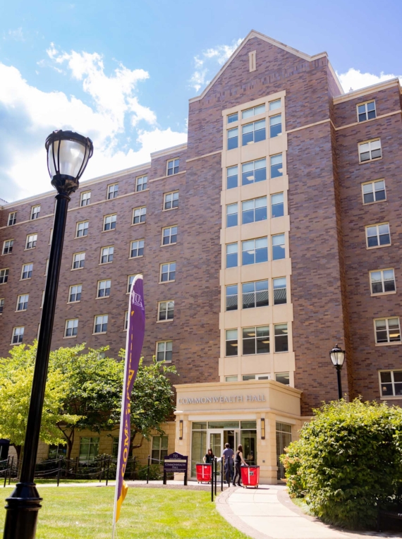 A multi-story brick building labeled Commonwealth Hall, part of West Chester University housing, features large windows and is surrounded by greenery and a sidewalk under a blue sky. A purple and yellow flag and two lamp posts stand in the foreground.