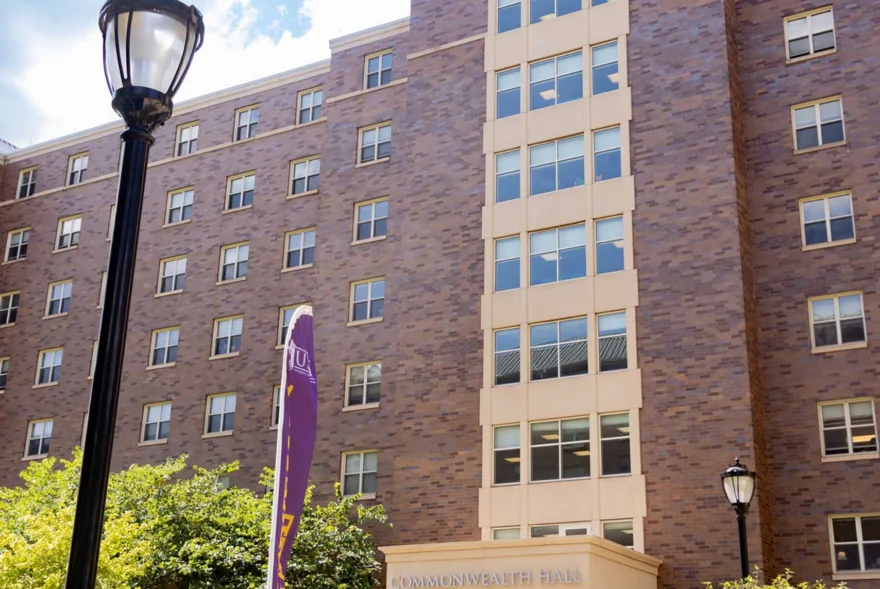 A multi-story brick building labeled Commonwealth Hall, part of West Chester University housing, features large windows and is surrounded by greenery and a sidewalk under a blue sky. A purple and yellow flag and two lamp posts stand in the foreground.
