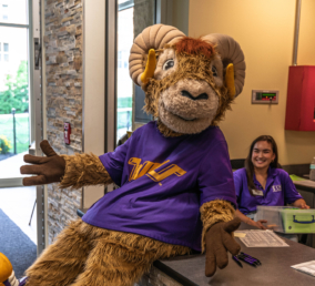A person in a ram mascot costume wearing a purple shirt sits on a counter with arms open. A smiling woman in a purple shirt, representing West Chester University housing, sits behind the counter with papers visible nearby in an indoor setting.