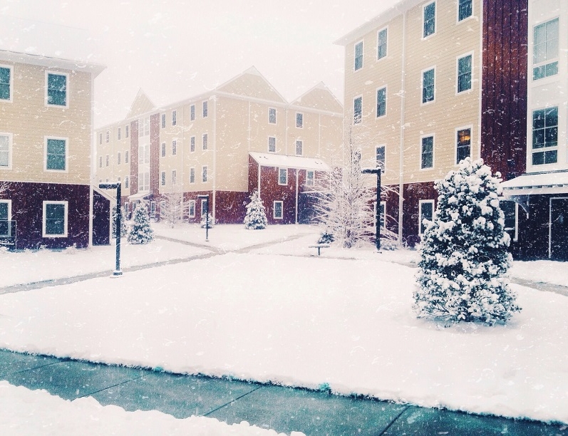 Snow-covered courtyard surrounded by multi-story beige and red apartment buildings, resembling West Chester University housing, with trees and bushes blanketed in snow. Snow is falling gently, creating a calm and wintry scene.